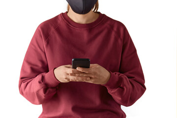 Cropped close up shot of a young european woman wearing face mask and burgundy sweatshirt holding a smartphone with both hands, typing a message, using application, isolated over white background.