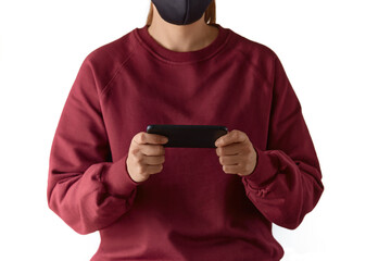 Cropped close up shot of a european woman wearing face mask and burgundy sweatshirt holding a smartphone horizontally with both hands, playing online, using application, isolated on white background.
