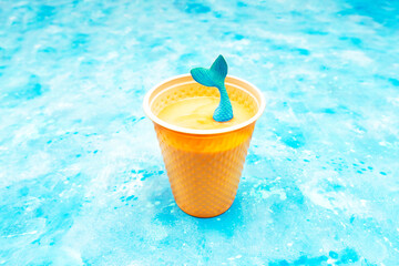 Toy mermaid tail sticks out of a plastic cup of cappuccino on a light blue background