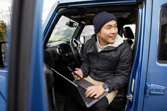 Happy man using smart phone and laptop on road trip in blue jeep