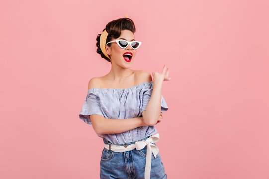 Enchanting pinup girl posing in sunglasses. Studio shot of slim young lady in striped blouse isolated on pink background.