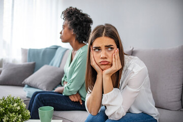 Two upset friends not talking to each other after fight on the sofa in sitting room at home. Two women friend sulking at each other, bad relationship concept.  Friendship, quarrel