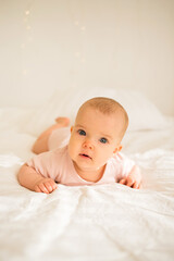 a little baby girl lies in a pink bodysuit on a white blanket on the bed and looks at the camera
