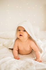 baby girl lies in a white towel with a hood on a white blanket on the bed and looks at the camera