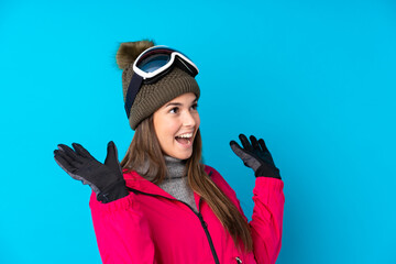 Teenager skier girl with snowboarding glasses over isolated blue background with surprise facial...