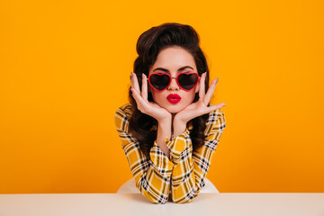 Pinup girl posing in heart shaped glasses. Lovely woman with bright makeup sitting on yellow background.
