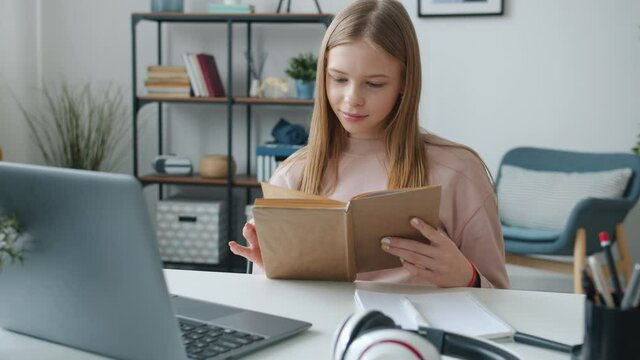 Female Student Is Reading Book Turning Pages Smiling Sitting At Desk In Apartment Alone Enjoying Literature. Childhood And Intelligence Concept.