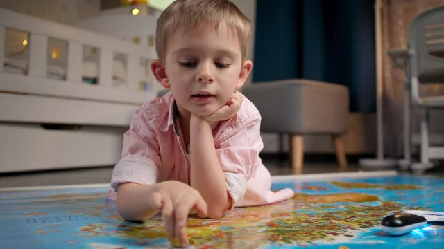 Cute smiling boy lying on floor and walking across countries with fingers on big world map. Concept of travel, tourism and child education. Kids exploration and discoveries.