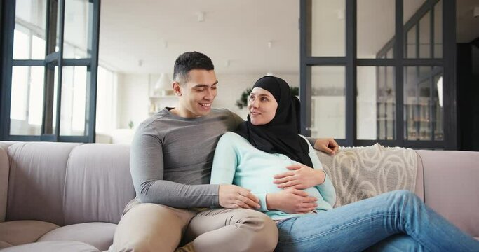 Happy Muslim pregnant woman in black hijab with smiling husband rests sitting on comfortable sofa in living room slow motion