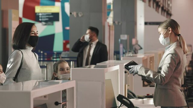 Medium shot of woman with kids checking-in at airport, handing passports to service agent and weighing luggage, while businessman having phone call in background, everybody wearing protective masks