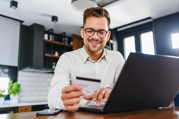 Young man holding credit card sitting in front of laptop computer at home paying for online order....