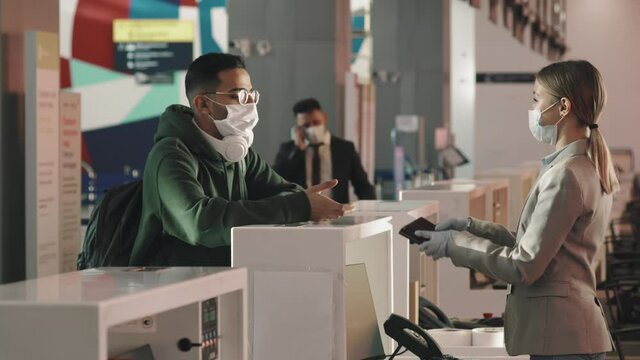 Medium shot of female airport service agent giving boarding pass to young man with backpack after passing passport control, while serious businessman talking on phone in background, all wearing masks