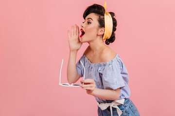 Pinup girl in striped blouse screaming to someone. Beautiful brown-haired woman with sunglasses posing on pink background.