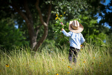 Little boy with cowboy hat in the field of grass and flowers, holding a flower bouquet he made by himself in his hand. 