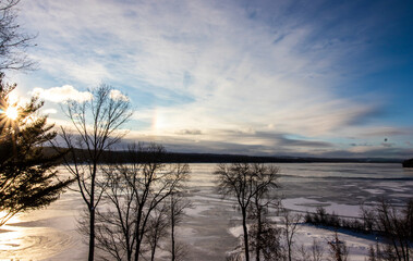 Frozen lake in winter with ice in Canada, dramatic sky and sunlight reflecting in the lake, bare trees in foreground, natural sunlight, horizontal image
