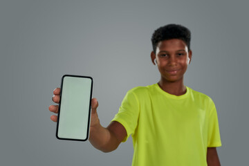 Happy teenage african boy wearing neon t shirt showing smartphone with blank screen at camera while standing against grey background