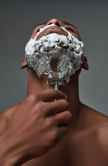 Close up of young african american man with foam applied on his face using steel razor for removing...