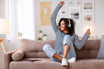 Happy ethnic woman listening to music and dancing on sofa