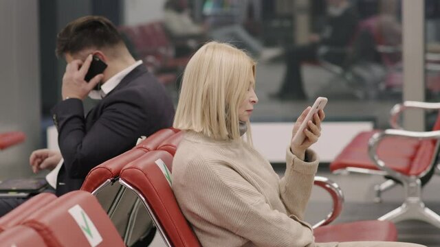 Medium slowmo side-view closeup of male and female strangers sitting back-to-back in departure lounge, businessman having phone call and woman surfing net, waiting for flight
