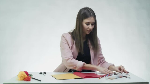 Attractive young woman uses pencil and ruler to draw lines on color paper at the table in a white studio