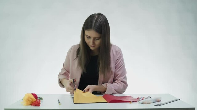 Attractive young woman uses scissors to cut lines of color paper at the table in a white studio