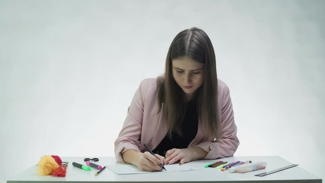Attractive young woman uses a black marker to draw something on on a white paper at the table in a white studio