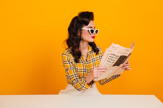 Elegant young woman reading newspaper. Studio shot of concentrated pinup girl posing on yellow background.