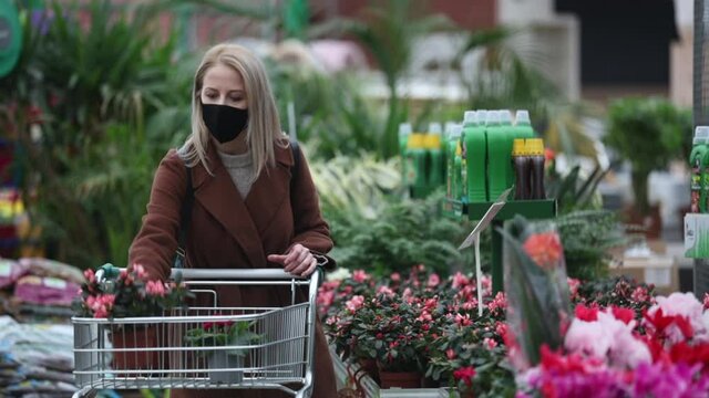 Blonde woman in face mask and coat is chooses flowers in a garden store