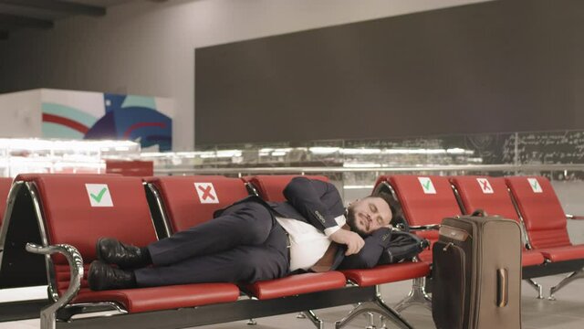 Slow-motion footage of business man in suit lying on seats in departure lounge sleeping while waiting for flight in empty airport