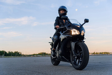 Fototapeta na wymiar Attractive girl in black leather jacket, pants and helmet on outdoors parking rides on stylish sports motorcycle at sunset.