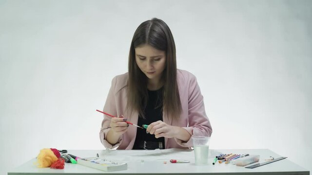 Attractive young woman draws with acrylic paints on a white paper at the table in a white studio