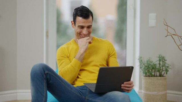 Relaxed cheerful Middle Eastern man laughing watching movie on tablet at home. Portrait of happy positive handsome guy enjoying weekend leisure indoors. Resting and relaxation concept. Slow motion.