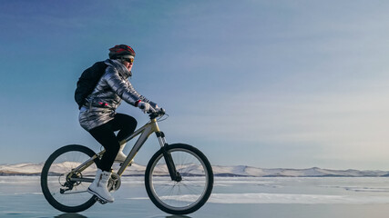 Woman is riding bicycle on the ice. Girl is dressed in a silvery down jacket, cycling backpack and helmet. Ice of the frozen Lake Baikal. Tires on bike are covered with spikes. Traveler is ride cycle.