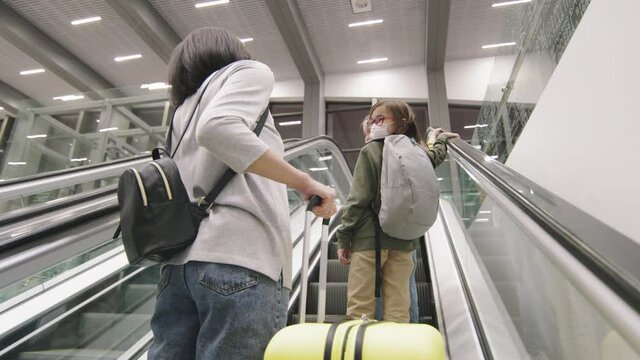 Low-angle back-view slowmo medium shot of woman with two little girl and big suitcase lifting on escalator in airport