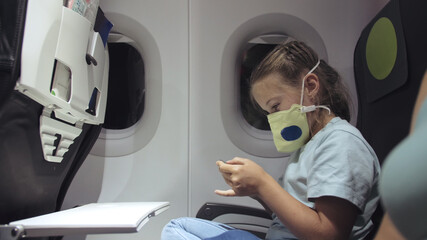 Fototapeta na wymiar Little girl caucasian at plane with wearing protective medical mask. Child baby tourist at aircraft with respirator play at smartphone mobile phone. Coronavirus epidemic sars-cov-2 covid-19 2019-ncov.