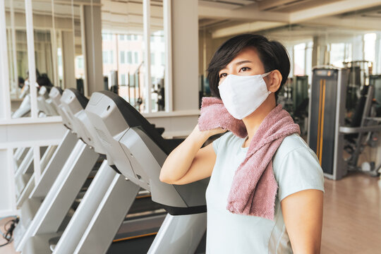 Exercising With A Face Mask - New Normal Lifestyle During Covid 19 Pandemic Concept. A Beautiful Asian Woman Wearing Cloth Mask While Working Out At The Gym With Fitness Equipments In The Background.