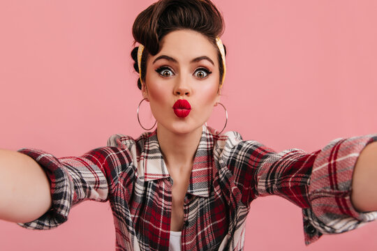 Jocund pinup girl posing in red checkered shirt. Amazed woman taking selfie with kissing face expression.