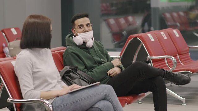 Medium shot of man and woman in protective masks chatting while sitting next to each other in departure lounge waiting for flight