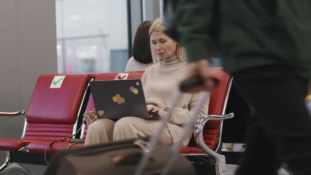 Medium slow-motion footage of adult caucasian woman working on laptop while waiting for flight in departure lounge with other passengers
