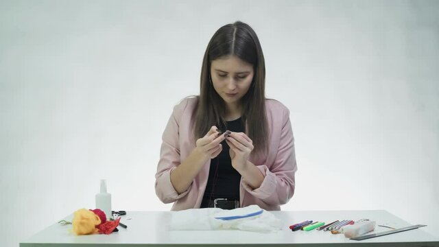 Attractive young woman threading a needle for cross stitching at the table in a white studio