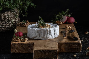 wooden board of camembert cheese garnished with walnuts , radish and herbs like rosemary and thyme on a dark background.
selective focus on the cheese.