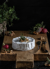 wooden board of camembert cheese garnished with walnuts , radish and herbs like rosemary and thyme on a dark background.
selective focus on the cheese.