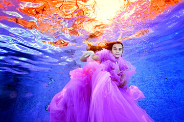 Fototapeta na wymiar A creative portrait of a young girl in a luxurious pink dress, who swims and plays underwater in the pool against the background of bright light on the surface of the water. Fashion photography.