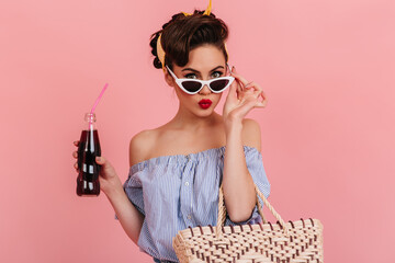 Attractive pinup woman drinking soda. Charming brunette girl in striped blouse touching sunglasses on pink background.
