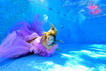 A young girl in a beautiful purple dress lies under the water at the bottom of the pool with her...