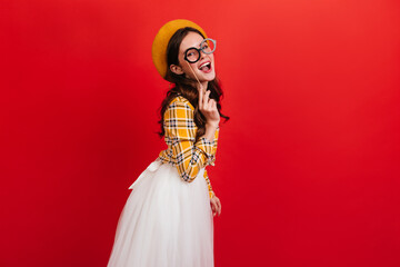 Indoor portrait of dark-haired girl in yellow shirt and white skirt. Lady in hat with smile posing with toy glasses