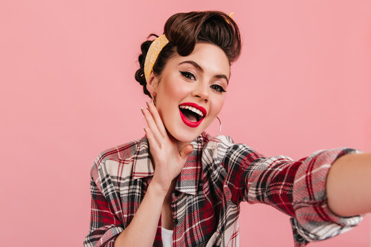 Amazed young lady in retro attire looking at camera. Winsome pinup girl taking selfie on pink background.