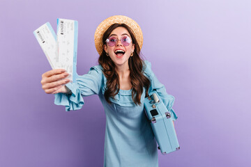 Portrait of young brunette in hat and glasses posing with suitcase on purple background. Woman in...