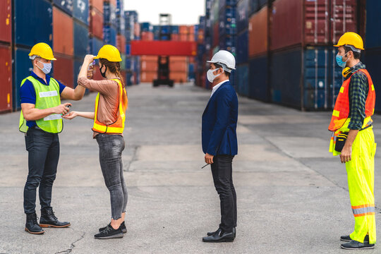 Professional of group cargo foreman in helmets standing and using infrared thermometer for checking body temperature staff fever before work in quarantine for coronavirus wearing protective mask