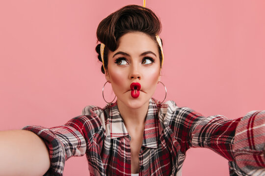 Cheerful pinup girl making funny faces. Fashionable brown-haired woman taking selfie on pink background.
